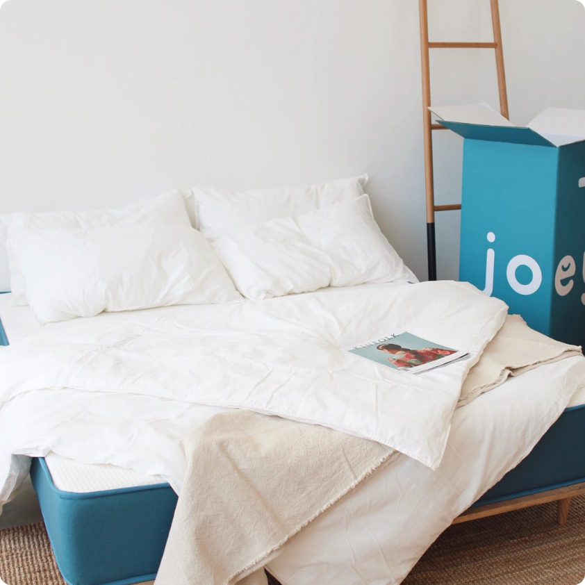 A Joey mattress with white bed sheets and a blue packaging box beside it in front of a decorative ladder