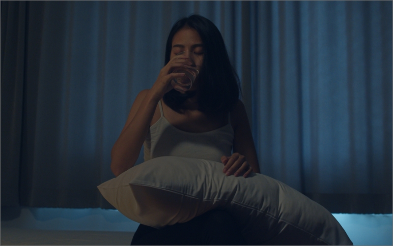Woman drinking water in a dark room while holding pillow on her lap