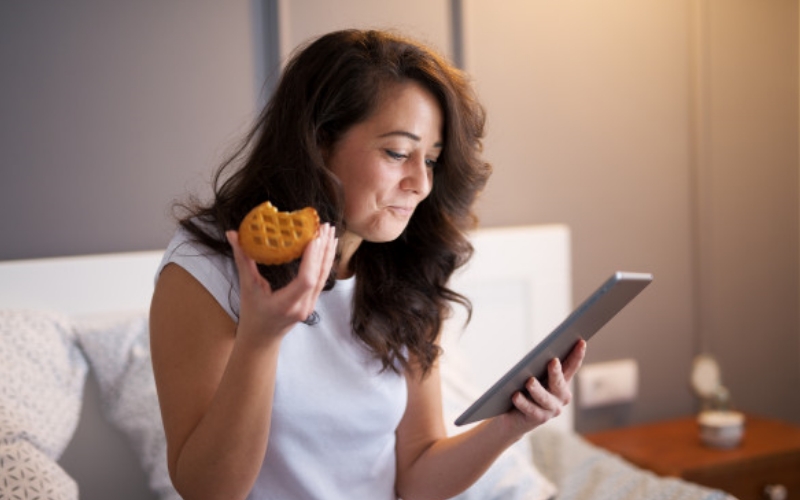 Woman eating a tart while looking at an iPad in bed